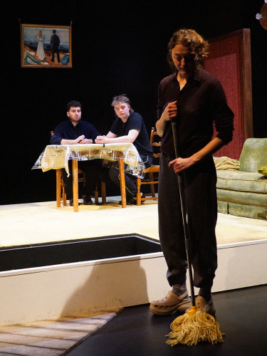 Eine Theaterprobe: Zwei Schauspieler sitzen im Hintergrund an einem Tisch. Im Vordergrund steht eine Schauspielerin mit einem Wischmop.