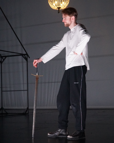 Ein Probenfoto: Ein Schauspieler steht lässig mit einem Schwert in der Hand auf der Bühne.