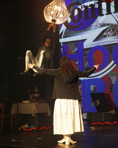 Ein Probenfoto: Ein Schauspieler schwingt an einem Kronleuchter. Eine Schauspielerin breitet ihm zugewandt die Arme aus.