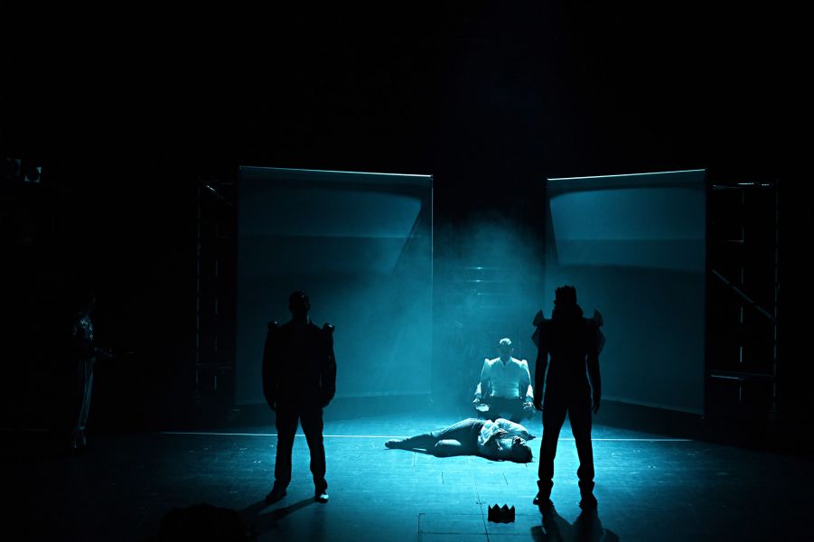 Das Foto zeigt im Vordergrund zwei dunkle Silhouetten. Dahinter liegt in einem blauen Licht eine Person auf dem Boden, eine weitere kniet über ihr.