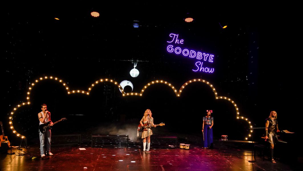 Das Bild zeigt vier Personen, die jeweils vor einem Mikron stehen und singen. Hinter ihnen befindet sich ein Lichterbogen und eine Leuchtschrift mit dem Titel The Goodbye Show