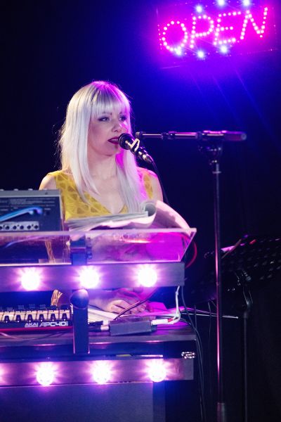 Das Foto zeigt eine Frau mit blondem Haar, die an einem Klavier sitzt und in ein Mikrofon singt.