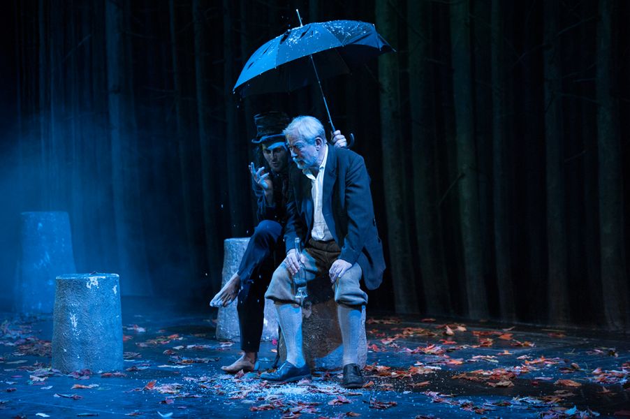 Auf dem Bild sitzen zwei Personen auf einem Baumstamm. An den älteren Mann lehnt sich der Boandlkramer und hält einen Schirm über Kaspar.