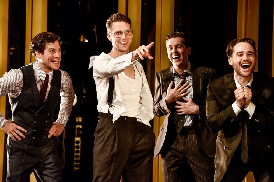 Das Foto zeigt vier Männer in Anzügen, die lachend in eine Richtung schauen. Vorne streckt ein Mann seine Hand nach vorne aus und zeigt auf etwas.