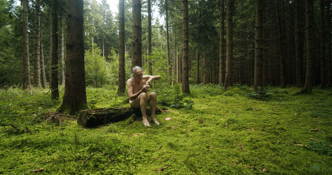 Das Foto zeigt einen alten Mann, der auf einem Baumstamm nackt sitzt und eine Dose öffnet. Um ihn herum ist grüner Wald.