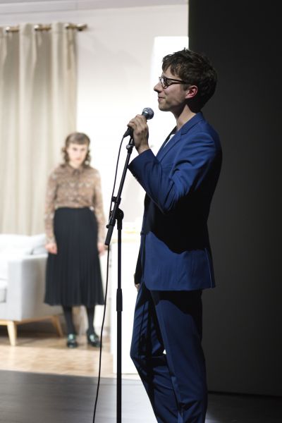 Das Bild zeigt einen Mann im blauen Anzug mit Mikrofon, weit hinter ihm steht Katharina Blum.