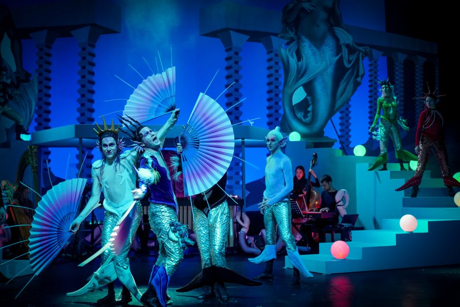 Das Bild zeigt ds Ensemble verteilt im Bühnenbild, einige tragen große Fächer und tanzen.
