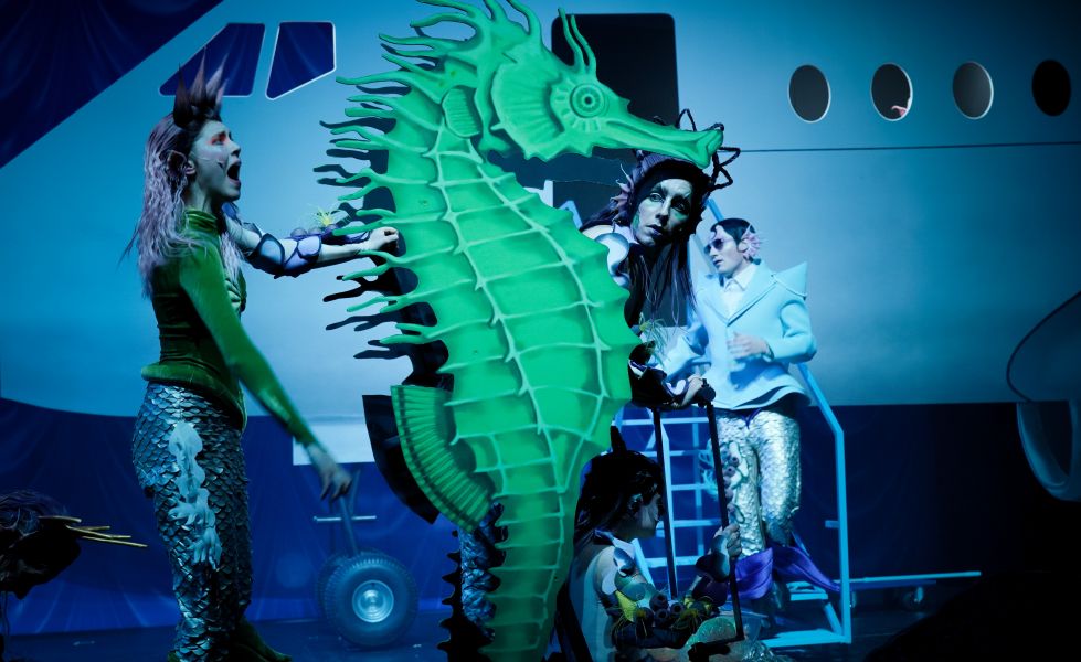Das Bild zeigt ein menschengroßes grünes Seepferdchen, das Ensemble darum verteilt und ein Flugzeug stehend im Hintergrund.