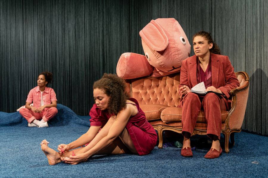 Das Bild zweigt zwei Darstellerinnen auf dem Boden und eine Darstellerin auf der Couch sitzend