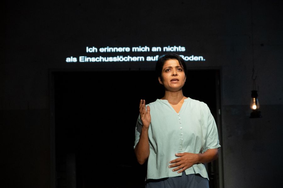 Ein Foto von Kavita Srinivasan vor einer Übertitelanzeige. Auf der steht: Ich erinnere mich an nichts als Einschusslöchern auf dem Boden.