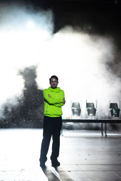 Ein Mann in neongrünen Sweatshirt steht auf einer Bühne, im Hintergrund ist Nebel.