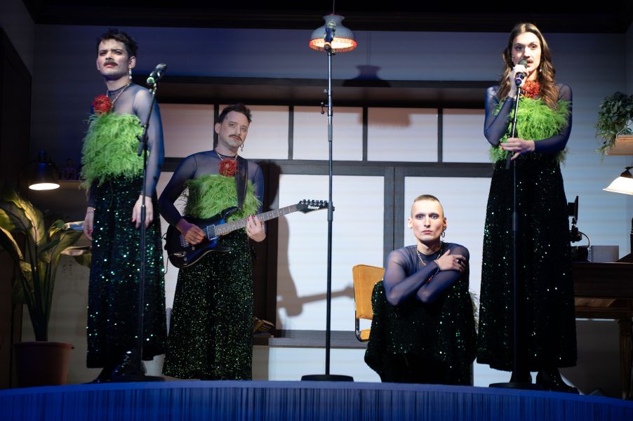 Drei Personen stehen auf der Bühne vor Mikrophonen, eine Person sitzt, eine weitere hält eine Gitarre. Sie tragen einen glitzerndes transparentes Shirt, ein knallgrünes Top darüber und einen langen schwarzen Rock.