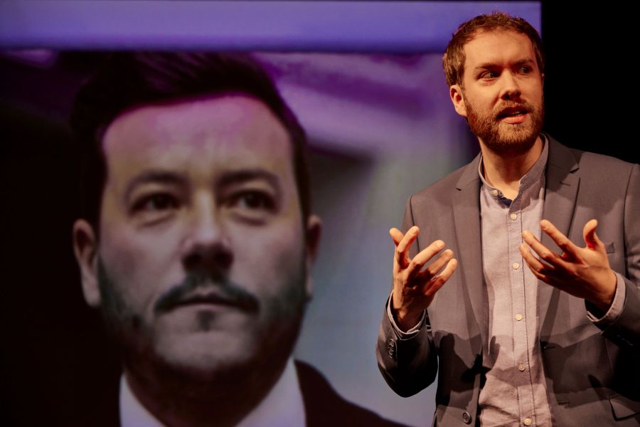 Person gestikuliert mit den Händen und steht vor einer Leinwand, auf der ein Portrait eines Mannes zu sehen ist.