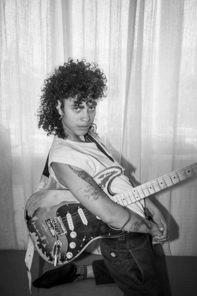 Eine Frau steht seitlich vor einem Vorhang und hält lässig eine E-Gitarre im rechten Arm.