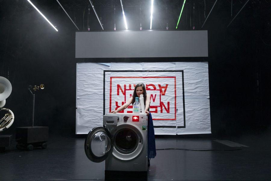 Eine Performerin steht hinter einer Waschmaschine, hinter ihr ein Schriftzug &quot;Never Again. Now.&quot;