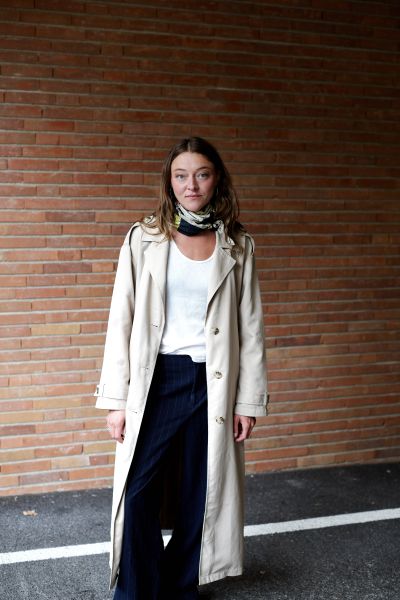 Lola Dockhorn im Trenchcoat vor einer Backsteinwand.